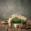 Three layered handmade soap bars with cream, green, and dark navy sections are arranged on a rustic wooden table, with a small bowl of green powder, cinnamon sticks, and small purple flowers as props against a textured gray background.